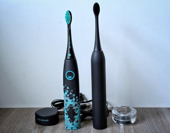 Die Happybrush Eco Vibe 3- und die Wondersmile PRO-Schallzahnbürsten inklusive Ladegeräte stehen nebeneinander auf einem Holztisch.
