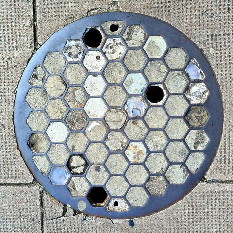 A round cover set into a pavement with hexagonal shaped holes outlibed by iron. Most of these hexagons have been filles in with concrete, but a few retain their original glass prisms.