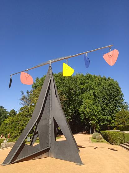 A 5 metre high frame of black metal rises like an arrowhead from a red-brown path. Balanced on its tip is a horizontal black bar, and from the bar are hanging, to the left of centre, a flat black form (far left) and a red form. Right of centre are a bright yellow form, a deep blue form and finally another brick red form. The forms sit above large trees in the distance, and stand against a brilliant blue sky