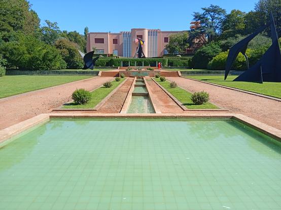 In the foreground, a sea-green tiled pool, square in form, of which we see three sides. From this extends a canal, ending where a short cascade enters it in the middle distance. Beyond this a flatly trimmed green hedge appears to support an enormous kinetic sculpture standing in front of a pinkish-red two-storey building in a modernist / art deco style. Paths of reddish stone flank the waterways, with lawns and trees to the far sides. Two more abstract sculptures of dark metal sit, one middle right, the other far left, on the grass