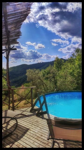 Portrait format photo of a wooden deck with a couple of deck chairs above a small circular blue pool. Below extends a landscape of hills and woods under a blue sky with some clouds. A red tiled roof is visible half way down the hill.