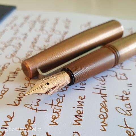 A picture of a Tactile Turn Gist fountain pen I in Copper with a Rose Gold plated nib unit from Bock
They lay on some handwriting in De Atramentis Document Ink Urban Sienna