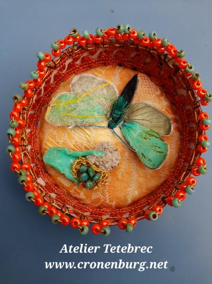 View from above into a richly decorated round box in shades of orange, green, gold and yellow. On a padded brocade fabric, the butterfly is pinned next to a decorative element. The inside edges are lined with braids. Around the edge is a handmade ribbon of perlage with floral elements in orange and green, made from the tiniest of limited edition rocailles and finest brass wire.