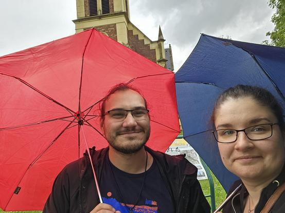 Selfie of me and Anna standing in front of Zábřeh Evangelic church both having umbrellas that cover almost all view of the church
