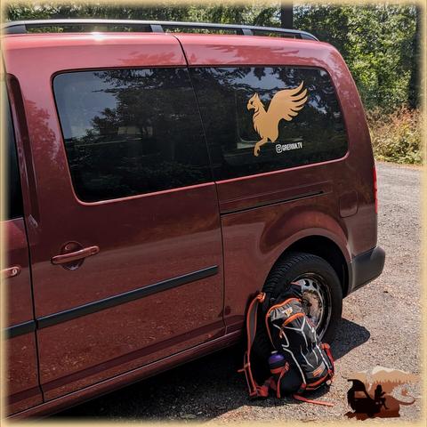 Foto auf die linke Schiebetür eines lavaroten Caddy Maxi mit goldener Beklebung. Der Wagen steht auf einem Kiesparkplatz im Wald. Am linken Hinterrad lehnt ein Wanderrucksack.