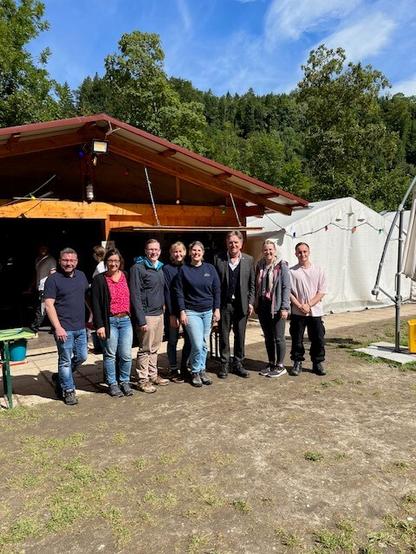 Minister Manne Lucha steht gemeinsam mit sieben weiteren Mitarbeitenden vom Zeltlager Benistobel in einer Reihe vor einer Holzhütte auf dem Zeltplatz.