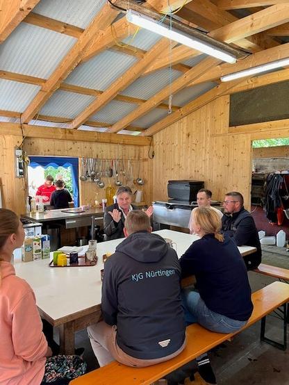 Minister Manne Lucha sitzt an einem Tisch in einer Küche. Mit ihm am Tisch sitzen fünf Mitarbeitende des Zeltlagers Benistobel. Er spricht und gestikuliert mit den Händen. Im Hintergrund sind durch ein Fenster Jugendliche zu sehen.