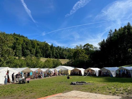 Man sieht eine Wiese, auf der mehrere große Zelte stehen. Zum Teil sind die Zelte geöffnet. In der Mitte der Wiese ist eine Grillstelle zu sehen. Im Hintergrund sind Bäume, vorne im Bild befindet sich ein Holztisch.