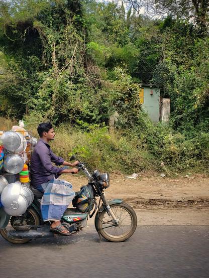 Man riding motorcycle on a road in Tamil Nadu loaded with pots and pans. Lush, green scenery in the background.