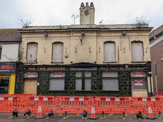 A derelict boarded up pub with buddleia sproutng and roadworks in front. The pub is called 'The Two Brewers'.