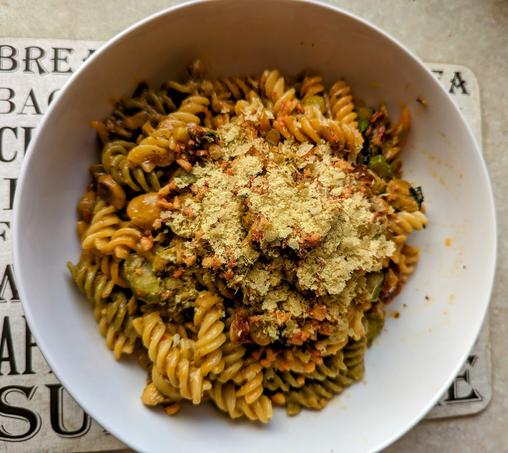 A white bowl of three coloured pasta (red, green and yellow) with chopped veggies and topped with nutritional yeast flakes.