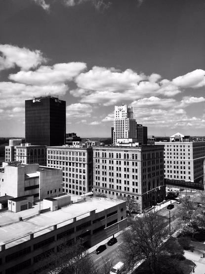 Black and white image of a block of downtown buildings viewed from the top floor of a city courthouse. The upper part of the image is a bright sky with puffy clouds, the middle of the image shows buildings of various heights in 1930s styles, including Art Deco, as well as a black, glass-clad tower on the left. The lower part of the image shows a street with cars and pedestrians and a 1990s parking deck.