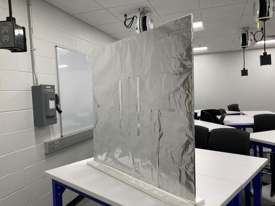 A COVID plexiglass protective screen covered in aluminum foil. There are two slits in the center of the screen, approximately 2 cm wide, at a distance of 20 cm apart. The screen is set up on a table in a lab classroom.