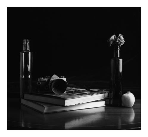 Black and white still life comprising two bottles, an Olympus OM2n camera, photo books and an apple.