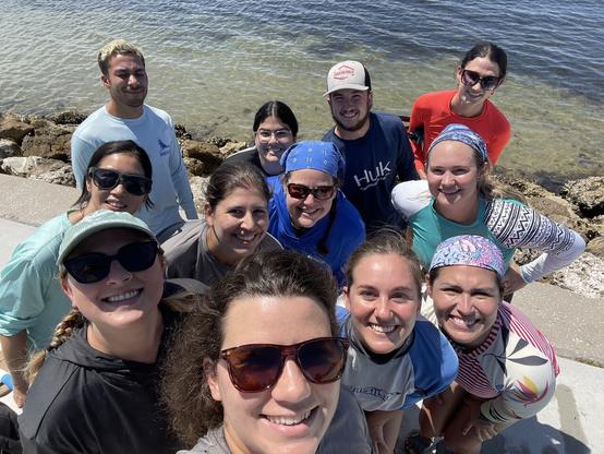 Selfie of a group of scientists standing in front of water containing a seagrass meadow - everyone looks very happy!
