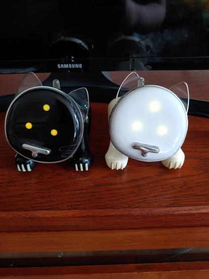 A black and white iCat are side-by-side on a table. Black one is on our left, white one on the right. Both tilted their heads to our left, ears up, and have 3 yellow LEDs lit up in a triangle. One on the upper-center, one on the bottom-left of that, and the other one on the bottom-right.