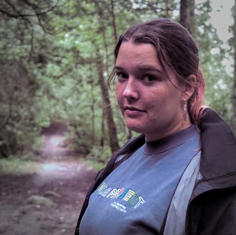 Girl facing camera wearing blue top and open light jacket over top in the foreground of a path through a forested area.