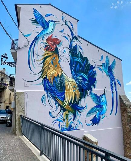 Streetartwall. On the upper external wall of a multi-storey house is sprayed/painted beautiful mural of a rooster with three birds. The house is visible until half. A bridge railing on the sidewalk in front of it shows that it must be on a mountain. The mural is mainly in blue tones and very fine and sweeping design. A large blue rooster with some yellow feathers and red head stands surrounded by blue short waves flying around like feathers. Around him fly three blue beautiful birds and complete the beautiful picture.