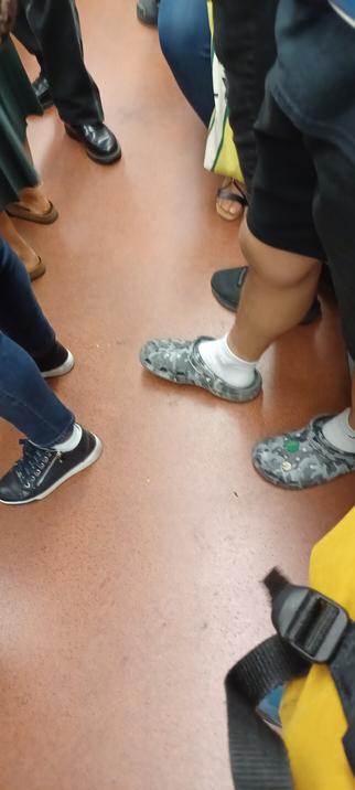 Photo des plusieurs paires de pieds dans une rame du métro parisien, l'une des paire est chaussée de croques en camouflage gris avec une Marguerite en plastique incrustée sur une face interieure