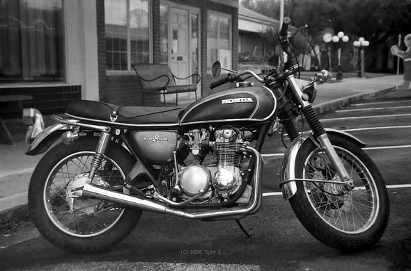 Monochrome black and white Foto of a Honda motorcycle on an empty street. Buildings in the background with striped parking spaces. Taken with an analog film camera.