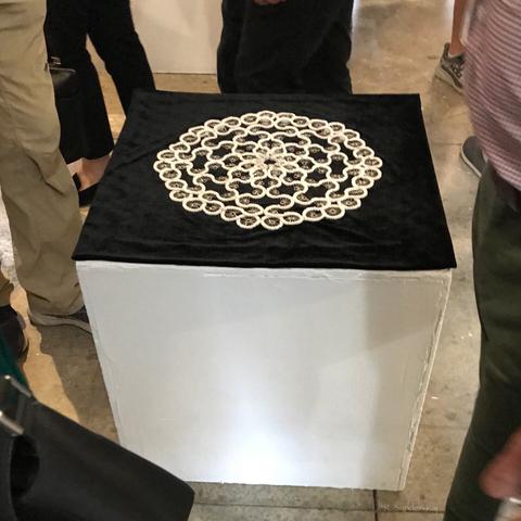 An 8-fold geometric pattern made of white cord and silver-coloured metal pieces on a black cloth on a white box.  It is seen at a downwards angle with a number of people standing around it.