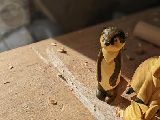 Loutre en bois debout bras le long du corps, pelage peint en marron, yeux et museau noir avec deux points blancs et un point gris. Posée sur un plan de travail. On voit partiellement deux autres sculptures (lapin et capybara)