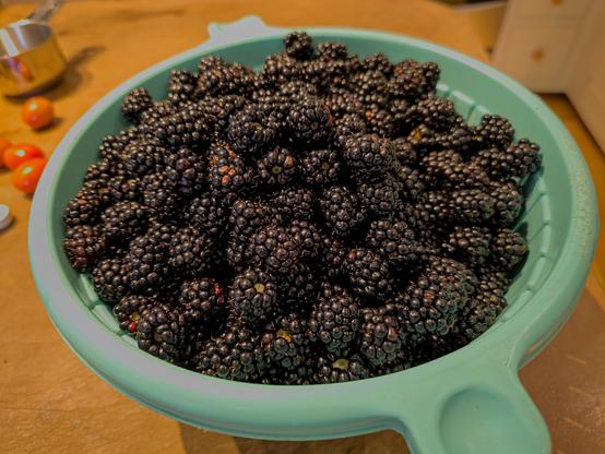 A green colander piled with blackberries is on a work surface.