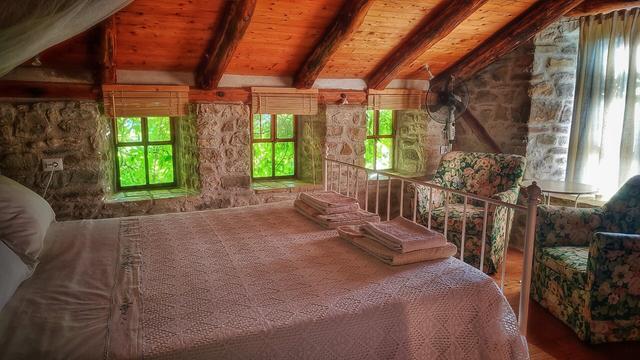 Photo of a bedroom with a double bed in the foreground under a sloping wooden roof with rustic round beams. To the right a tall window with sunlight shining through the curtains and in the far wall three small low 4-paned windows with green foliage outside.