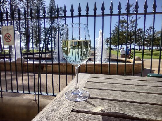 A glass of semillon wine sitting on a table outside with a fountain behind and a beautiful blue sky.