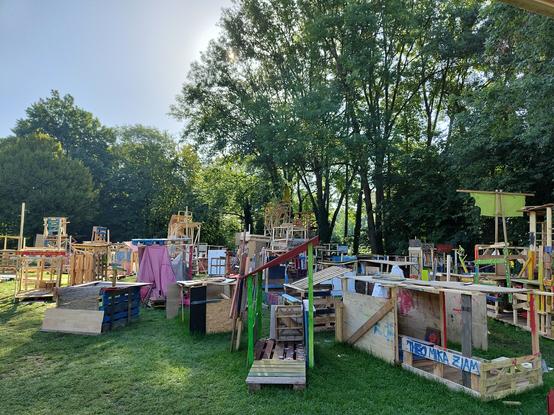 Zahlreiche von Kindern erbaute Konstrukte aus Holz, auf einer Wiese, über dem Darmbach, um Bäume herum.