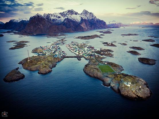 Aerial shot. Two elongated rocky islands surrounded by other small islands in the center, a snow-covered mountain range directly bordering the sea in the background.

The two main islands are built up with houses in the back part forming a small village. On the right island, a soccer field can be seen in the front part.