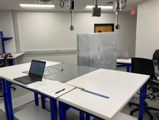 A receiver software defined radio antenna, magnetically attached to a steel ground plane. The antenna is behind a double-slit made of aluminum foil and is hooked up to a laptop running SDR software. There is a lab book to the right of the laptop for data logging.