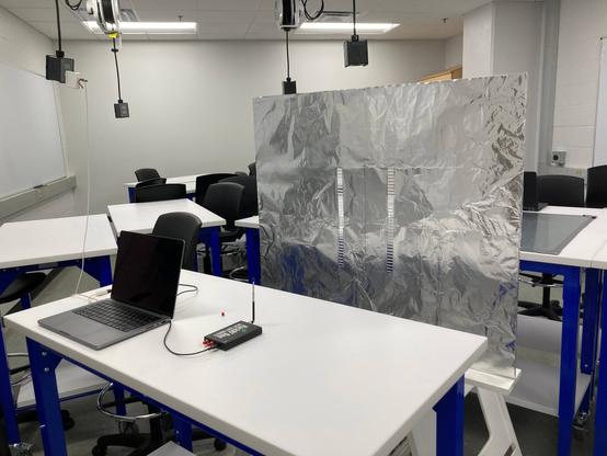 A lab setup of a transmitting antenna attached to a software-defined radio that is connected to a laptop computer. The antenna is in front of a double slit made of aluminum foil.