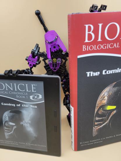 A photo of a robotic, humanoid, purple-and-black Lego/Bionicle creation, holding up a physical book and a Kindle displaying the same book on its screen. The book is "Biological Chronicle Book 1: The Coming of the Toa."