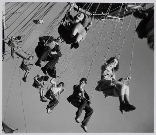 Photographie en noir et blanc en contre-plongée.
Des personnes radieuses voltigent dans les airs : elles sont sur un manège de chaises volantes. 
On ne voit pas le manège lui-même. 
L’image évoque la légèreté et l’insouciance.