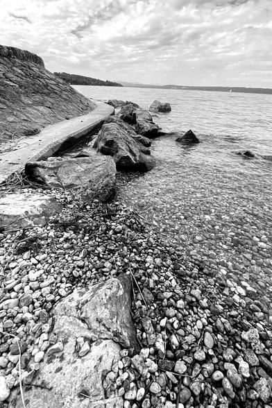 Black and white photo showing a rocky beach in the foregeound, Ammersee in Germany in the background.