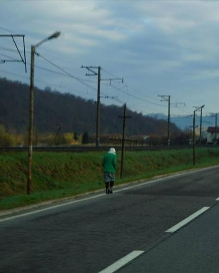 Masha Reva, road series made when the artist left Uzhhorod (Ukraine) for Berlin, April 2022 // A stooped lady in green walks along the road. On the lady's head is a light scarf. The lady is taken from behind. Behind the track, iron tracks, power lines and a dark forest are visible. The mountains are blue in the distance. // Маша Рева, роуд-серия, сделанная при отъезде из Ужгорода в Берлин, конец апреля 2022 года // Сутулая дама в зелёном идёт вдоль трассы. На голове дамы светлый платок. Дама снята со спины. За трассой видны железные пути, линии электропередач и тёмный лес. Вдали синеют горы.