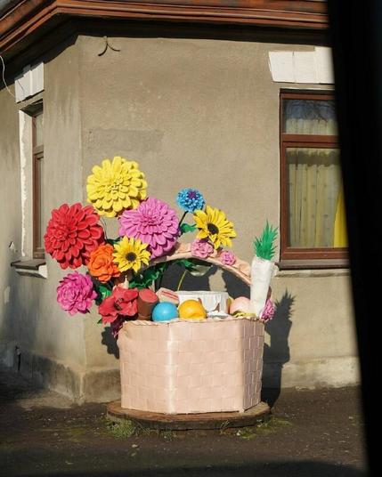 Masha Reva, road series made when the artist left Uzhhorod (Ukraine) for Berlin, April 2022 // Decorative basket with giant artificial flowers and fake eggs in different colours. The basket is installed on the corner of a one-story gray brick house. // Маша Рева, роуд-серия, сделанная при отъезде из Ужгорода в Берлин, конец апреля 2022 года // Декоративная корзина с гигантскими искусственными цветами и ненастоящими яйцами разных цветов. Корзина установлена на углу одноэтажного кирпичного дома серого цвета.