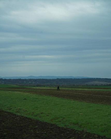Masha Reva, road series made when the artist left Uzhhorod (Ukraine) for Berlin, April 2022 // In the center of the photo, two people are walking across a field. Otherwise, it is a rural landscape with a blue cloudy sky, four electrical wires hanging between poles that are not included in the frame, a green and brown field, a village, a forest and mountains in the distance. // Маша Рева, роуд-серия, сделанная при отъезде из Ужгорода в Берлин, конец апреля 2022 года // В центре фотографии два человека идут по полю. В остальном это сельский пейзаж с синим облачным небом, четырьмя электрическими проводами, висящими между столбами, не вошедшими в кадр, зелёным и коричневым полем, селом, лесом и горами вдали.