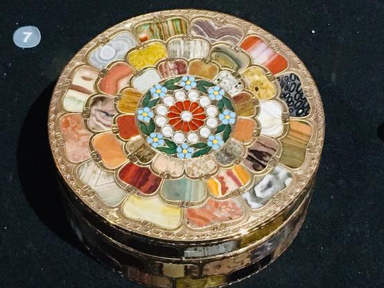 Photographie en couleur. 
Petite boîte ronde pour tabac à priser. 
Mosaïque de pierres dures multicolores et d’or : agate, onyx, jaspe, cornaline, améthyste… 
La mosaïque forme un décor floral, chaque pierre représentant un pétale.