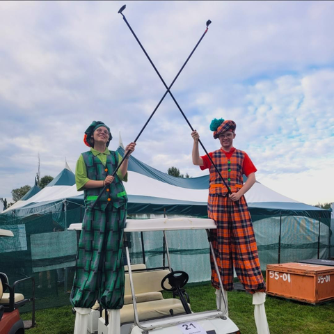 Two people on stilts in old time Scottish golfing outfits. On the left green plaid and the on the right orange plaid. They’re holding long poles crossed between them over their heads. The poles are for retrieving golf balls.