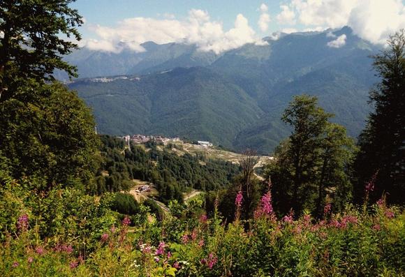 Mountain landscape. overlooking the valley.  buildings are visible, from a great distance they seem tiny. majestic mountains and clouds above them are also visible. trees, grass, flowers are visible in the foreground