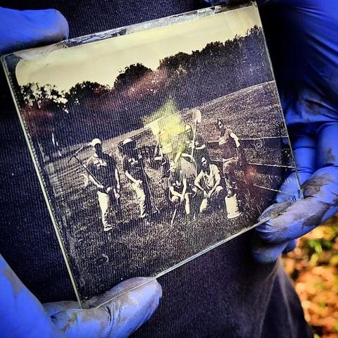 A wet plate photo, held by two gloved hands, shows a blurry image of a group of Archaeologists holding tools.