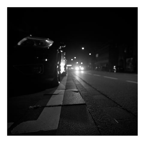 Aufnahme einer Straßenszene bei Nacht. Das linke Drittel des Bildes zeigt ein geparktes dunkles Auto auf dem Gehweg. Einige Gehwegplatten zeigen Risse oder sind uneben. Eine der Platten wirft durch den Scheinwerfer eines auf der Straße fahrenden Autos einen langen Schatten. In der rechten unteren Ecke liegt eine Zigarette am Bordstein.