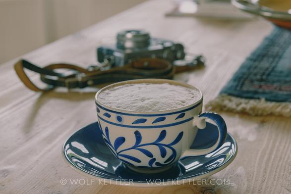 A coffee cup and, behind and blurred, vintage looking camera on a wooden table, processed to look vintage/retro. Very kitschy!
