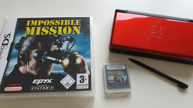 Auf dem Tisch liegend: 
Coveransicht der Spieleverpackung "Impossible Mission" für Nintendo DS, das Modul des Spieles, eine Nintendo DS Light in Crimson Red, der Stylus zur Nintendo DS.