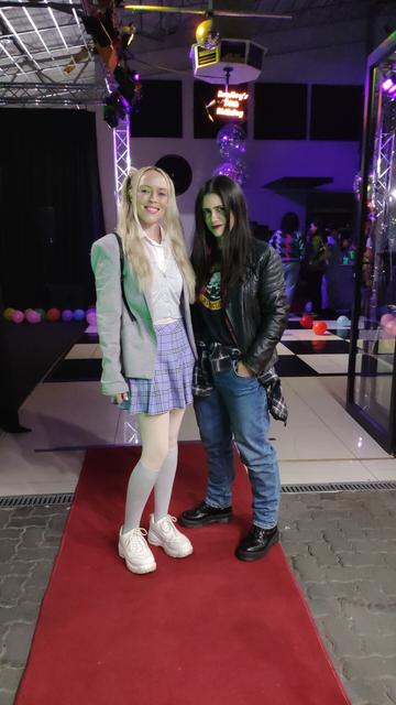 Photo from a 90s themed birthday party with a a blonde in preppy Clueless dress, and brunette in the grunge style, with docs, jeans, leather jacket and plaid.