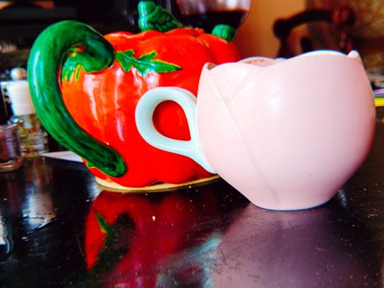 Une théière tomate de trois quarts dos : la Hanse, capuchon et le bec sont des feuilles vertes et le corps une tomate très charnue rouge, esthétique vieille vaisselle kitsch. En céramique sur un dessous de verre de bar. Deva 't de trouve Une tasse rose pastel en forme de tulipe à la Hanse vert pastel comme une feuille en esthétique minimamiste mignonne asiatique en céramique. Autour c'est cradeau désolé