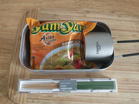In einem rechteckigen Campinggeschirr aus Alu (Messtin) liegen ein Paket Instant-Nudelsuppe und ein kleiner Spirituskocher. Darunter ein paar Essstäbchen