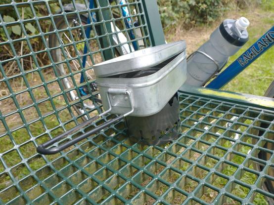 Ein Messtin/Alutopf steht auf einem Topfständer mit Windschutz, darin kocht Wasser auf. aufgebaut ist alles auf einer Parkbank.